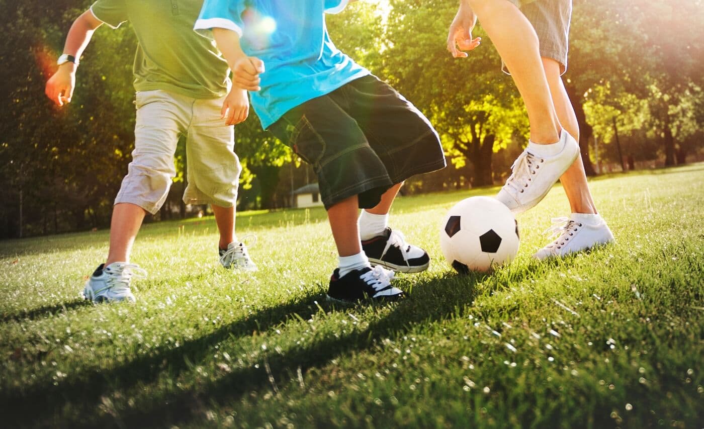 Kids playing in the grass with a soccer ball, basketball, football, and a Bag of Bases set up around them